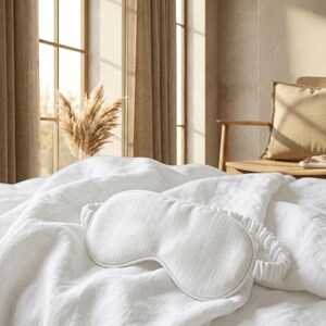 May include: A white sleep mask with a textured fabric surface and a matching gathered elastic strap. The mask features a contoured shape designed to cover the eyes, with clean stitched edges and a consistent off-white color throughout the piece.