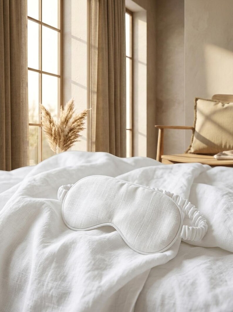 May include: A white sleep mask with a textured fabric surface and a matching gathered elastic strap. The mask features a contoured shape designed to cover the eyes, with clean stitched edges and a consistent off-white color throughout the piece.