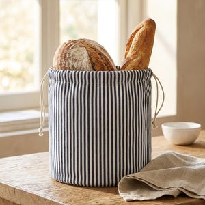 Könnte beinhalten: Ein zylindrischer Brotsack aus Stoff mit einem vertikalen Streifenmuster in Marineblau und Weiß und einem Kordelzug am oberen Rand. Der Sack ist mit einem runden Laib Brot und einem langen Baguette gefüllt, die aufrecht darin stehen.