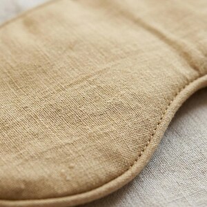May include: A tan sleep mask featuring a visible woven fabric texture. The mask has rounded edges and is constructed with a stitched border that follows the curved contour of the piece, highlighting the natural fiber weave of the material.