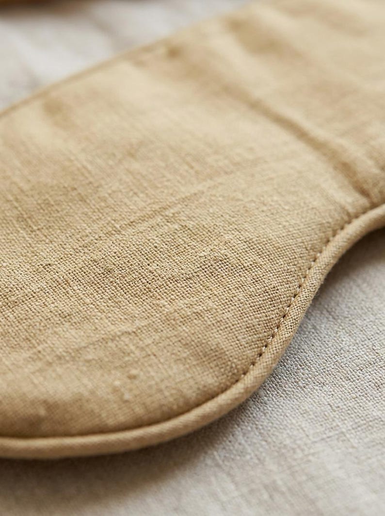 May include: A tan sleep mask featuring a visible woven fabric texture. The mask has rounded edges and is constructed with a stitched border that follows the curved contour of the piece, highlighting the natural fiber weave of the material.