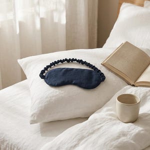 May include: A dark navy blue sleep mask featuring a smooth, contoured front panel and a gathered, elasticized fabric strap. The mask is designed to cover the eyes for light blocking and rests on a white pillow next to an open book and a ceramic cup.