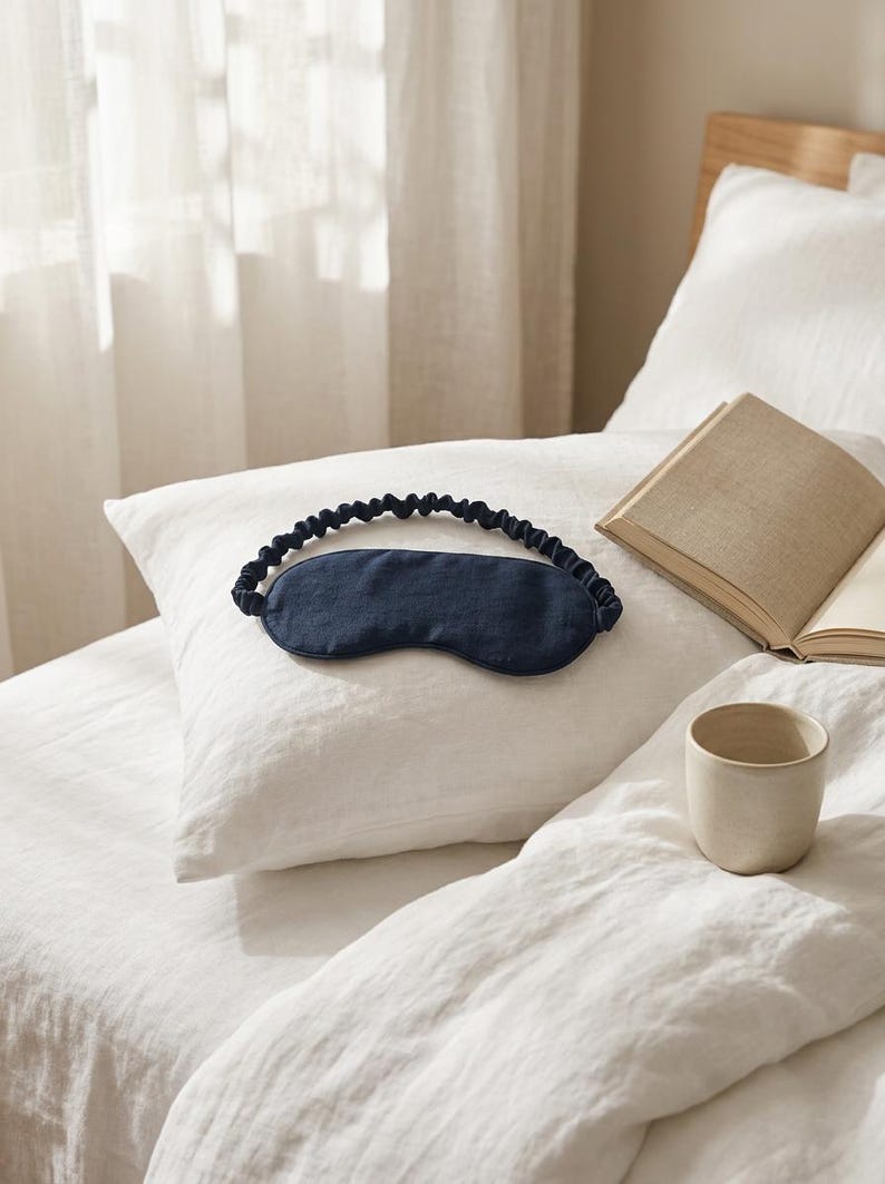 May include: A dark navy blue sleep mask featuring a smooth, contoured front panel and a gathered, elasticized fabric strap. The mask is designed to cover the eyes for light blocking and rests on a white pillow next to an open book and a ceramic cup.