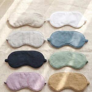 May include: A collection of eight sleep masks arranged in two columns. The masks feature various solid colors including beige, white, light gray, blue, dark navy, sage green, lavender, and mustard yellow, each with a simple contoured shape.