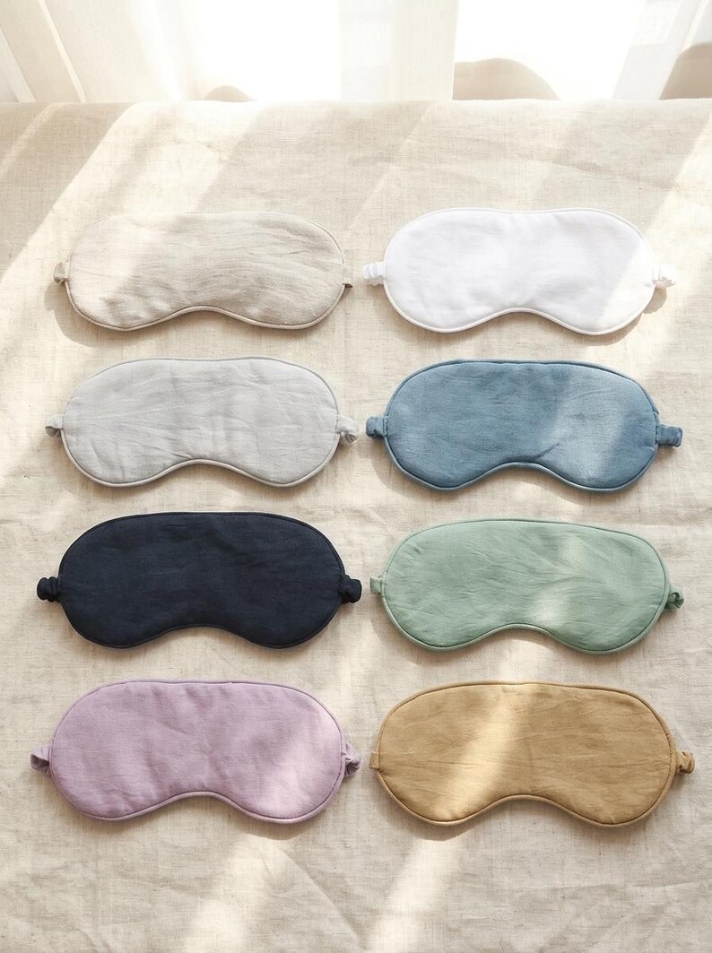 May include: A collection of eight sleep masks arranged in two columns. The masks feature various solid colors including beige, white, light gray, blue, dark navy, sage green, lavender, and mustard yellow, each with a simple contoured shape.