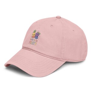 Könnte beinhalten: Eine hellrosa Baseballkappe mit einer Grafik aus einer lächelnden Ananas, Weintrauben und einer Blaubeere. Unter der Fruchtillustration steht der Text "PROUD TO BE FRUITY" in bunten, blockartigen Buchstaben. Die Kappe hat einen gebogenen Schirm.