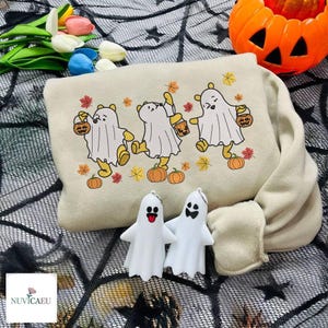 May include: A beige sweatshirt featuring an embroidered design of three yellow bears dressed as ghosts, each holding a small orange pumpkin bucket. The design is surrounded by scattered orange, red, and yellow autumn leaves. Two white plastic ghost figurines are placed in front.