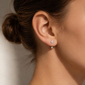 May include: A double-ended earring featuring a small, clear, round faceted stone stud on the front of the earlobe and a smooth, polished silver-toned spherical bead positioned directly beneath the lobe, creating a suspended, layered effect.