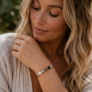 May include: A beaded bracelet featuring a sequence of smooth, round stones in shades of white, light blue, translucent grey, and black, separated by small silver-toned spacers and textured, dark grey, porous, square-shaped accent beads.