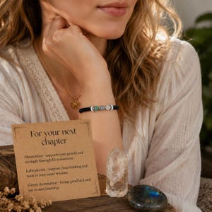 May include: A black cord bracelet featuring three round gemstone beads in green, blue, and white, accented by two silver-toned crescent moon charms. A tan card with the text For your next chapter, a clear quartz crystal point, and a polished labradorite stone.