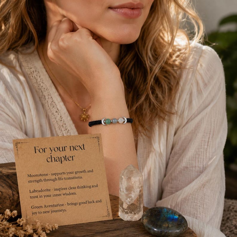 May include: A black cord bracelet featuring three round gemstone beads in green, blue, and white, accented by two silver-toned crescent moon charms. A tan card with the text For your next chapter, a clear quartz crystal point, and a polished labradorite stone.