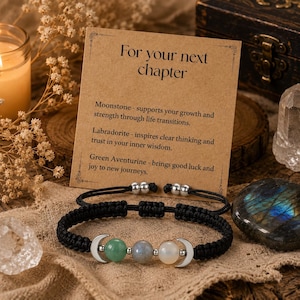 May include: A black cord bracelet featuring three round gemstone beads in green, grey, and white, flanked by two silver-toned crescent moon charms. The bracelet is displayed on a tan card with the text For your next chapter and descriptions of Moonstone, Labradorite, and Green Aventurine.