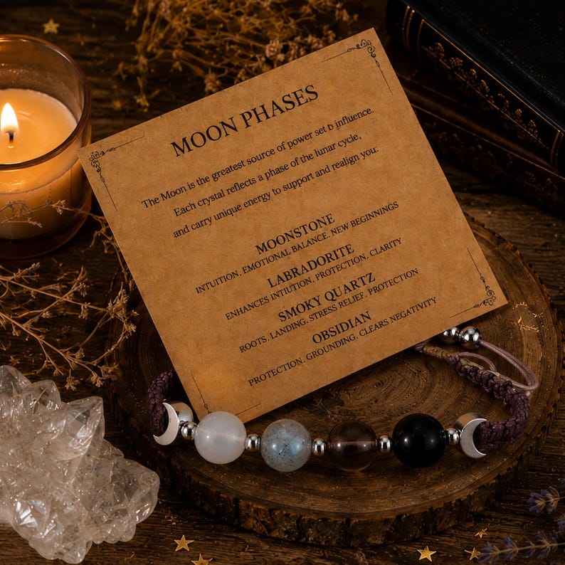May include: A bracelet featuring four spherical gemstones: white moonstone, grey labradorite, translucent smoky quartz, and black obsidian. The beads are separated by silver-toned spacers and crescent moon charms on a dark brown cord.