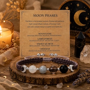 May include: A brown woven cord bracelet featuring four spherical beads in white, grey, brown, and black, accented by two silver-toned crescent moon charms. The bracelet is displayed in front of a tan card printed with the text MOON PHASES.