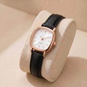 Peut inclure: Une montre à quartz en or rose avec un cadran carré et blanc. La montre a des bracelets en cuir noir. La montre est présentée sur une surface beige et incurvée.