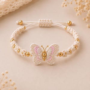 May include: A macrame bracelet featuring a central butterfly charm with cream and light pink thread details. The band is constructed from cream-colored cord, accented with small, round, polished gold-tone metal beads and an adjustable sliding knot.