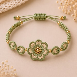 Puede incluir: Una pulsera de macramé con un motivo floral central en hilo verde salvia y crema, acentuado con pequeñas cuentas metálicas doradas. La banda consiste en un cordón trenzado verde con elementos en forma de hoja y un cierre de nudo corredizo.