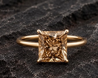 May include: A gold ring featuring a large, square-cut, champagne-colored gemstone set in a four-prong setting. The band is smooth and polished, with the text Verve Jewel visible in the background. The gemstone displays a faceted, brilliant cut.