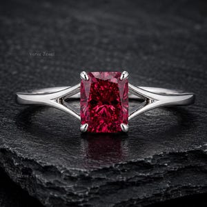 May include: A silver-toned ring featuring a cushion-cut, deep red gemstone held by four prongs. The band has a split-shank design that tapers toward the center stone. The text Verve Jewel is visible in small, faint lettering on the surface.
