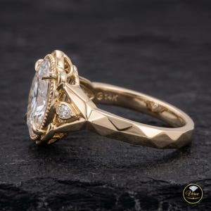May include: A 14K yellow gold ring featuring a large oval-cut centre stone surrounded by a beaded halo setting. The band has a faceted, geometric design with small pear-shaped accent stones on the sides. The interior is stamped with 14K.