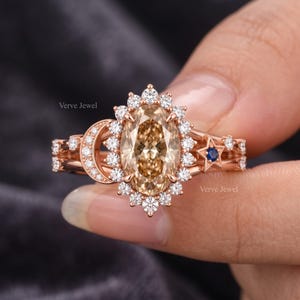 May include: A rose gold ring featuring a central oval-cut champagne-coloured gemstone surrounded by a halo of small white stones. The band includes a crescent moon shape on one side and a star with a small blue stone on the other. Text reads Verve Jewel.