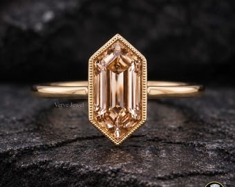 May include: A gold ring featuring a central hexagonal-cut champagne-colored gemstone set within a milgrain-detailed bezel. The band is smooth and polished gold, and the setting is oriented vertically. The text Verve Jewel is visible on the left.