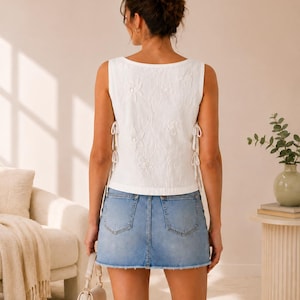May include: A white sleeveless top featuring an embroidered floral pattern and side lace-up ties, paired with a light blue denim mini skirt with a raw hem. The top has a square neckline and the skirt includes two back pockets and a classic button closure.