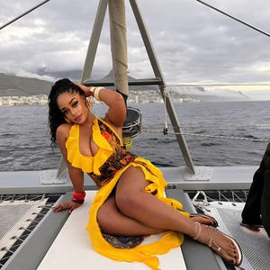 Pu&ograve; includere: Una donna indossa un abito giallo con scollatura a balze e corpetto fantasia, posa su una barca a vela. L'abito ha una gonna fluida. Indossa sandali e bracciali dorati. Lo sfondo mostra acqua e un cielo nuvoloso.