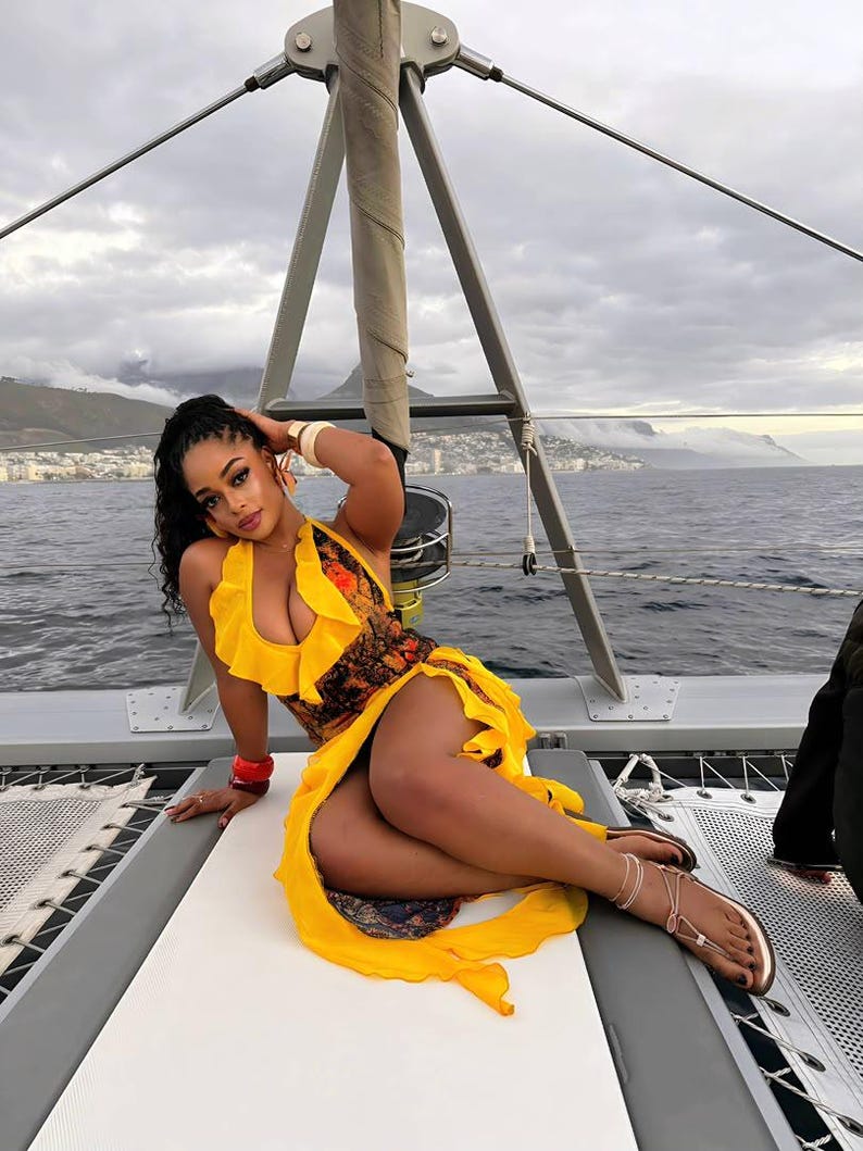 Pu&ograve; includere: Una donna indossa un abito giallo con scollatura a balze e corpetto fantasia, posa su una barca a vela. L'abito ha una gonna fluida. Indossa sandali e bracciali dorati. Lo sfondo mostra acqua e un cielo nuvoloso.