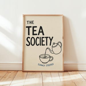 Könnte beinhalten: Ein rechteckiger gerahmter Kunstdruck auf cremefarbenem Hintergrund mit dem schwarzen Text THE TEA SOCIETY. Unter dem Text befindet sich eine schwarze Strichzeichnung einer Teekanne, die Flüssigkeit in eine Tasse mit einem Blatt gießt. Darunter steht der blaue Text SLOWLY STEEPED.