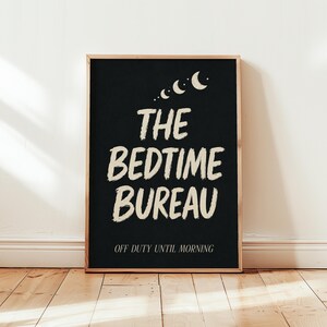 May include: A rectangular wall art print in a wooden frame featuring a black background with cream-colored text that reads THE BEDTIME BUREAU and OFF DUTY UNTIL MORNING, accompanied by a series of four crescent moon icons at the top center.