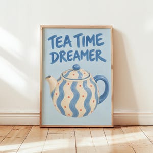 May include: A rectangular wall art print featuring a light blue background with the text TEA TIME DREAMER in bold blue letters. Below the text is an illustration of a cream-coloured teapot with blue vertical wavy stripes and red dots.