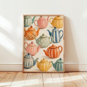 Op de afbeelding: Een ingelijste kunstdruk met een verzameling van elf keramische theepotten in diverse kleuren en patronen, waaronder strepen, stippen en bloemmotieven. De theepotten zijn in een rasterachtige compositie op een crèmekleurige achtergrond geplaatst.