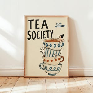 Op de afbeelding: Een rechthoekige wandkunstprint met de tekst TEA SOCIETY en SLOW AFTERNOON in zwart en blauw. De illustratie toont een stapel van vier keramische theekopjes, met een persoon die ontspant in het bovenste kopje, versierd met blauwe, rode en witte patronen.
