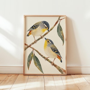 May include: A framed art print featuring two small birds perched on thin branches with green leaves. The birds have yellow chests, grey wings with white spots, and black-and-white patterned heads. The background is a solid light cream color.