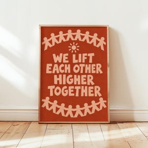 Puede incluir: Un póster rectangular con fondo terracota que presenta un icono de sol en color melocotón claro y el texto WE LIFT EACH OTHER HIGHER TOGETHER en una fuente redondeada, enmarcado por dos filas de figuras de papel estilizadas tomadas.