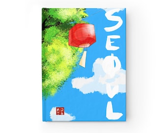 Puede incluir: Un diario de tapa dura con una ilustración digital de una linterna roja colgando contra un cielo azul brillante con nubes blancas y follaje verde. La palabra SEOUL está escrita en letras blancas grandes a la derecha, con un pequeño logo cuadrado rojo.