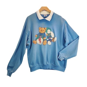 May include: A light blue sweatshirt with a white collar and a design featuring two embroidered teddy bears. One bear wears overalls and the other a bonnet. A heart-shaped gift is between them.