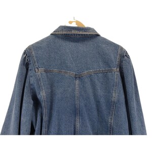 May include: Rear view of a blue denim jacket suspended on a wooden hanger. The jacket features a traditional collar, button fastenings, and a fitted design. The denim is a medium wash, with brown stitching providing contrast.