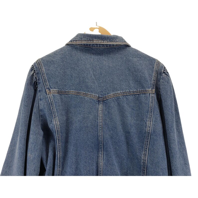 May include: Rear view of a blue denim jacket suspended on a wooden hanger. The jacket features a traditional collar, button fastenings, and a fitted design. The denim is a medium wash, with brown stitching providing contrast.