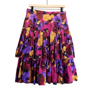 May include: A tiered midi skirt featuring a vibrant floral pattern in shades of magenta, purple, and golden yellow. The skirt has a fitted waistband and two distinct horizontal ruffled layers that create a voluminous, gathered silhouette.