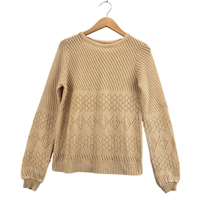 Puede incluir: Suéter de color beige con cuello redondo y mangas largas. El suéter presenta un diseño texturizado con recortes en forma de diamante en el pecho y las mangas. Cuelga de una percha de madera sobre un fondo blanco.
