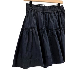 Puede incluir: Falda escalonada azul marino con cintura fruncida, colgada de una percha de madera. La falda tiene una silueta acampanada y est&aacute; hecha de una tela ligera. La falda es una pieza vers&aacute;til para cualquier guardarropa.