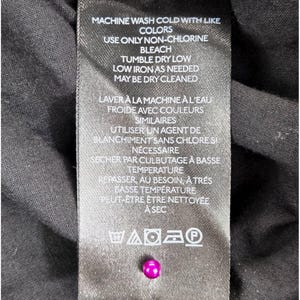 Puede incluir: Una etiqueta de ropa negra con texto blanco que detalla las instrucciones de lavado. La etiqueta incluye instrucciones en ingl&eacute;s y franc&eacute;s, con s&iacute;mbolos para lavar a m&aacute;quina, blanquear, secar y planchar. Una peque&ntilde;a cuenta rosa brillante est&aacute; unida a la etiqueta.