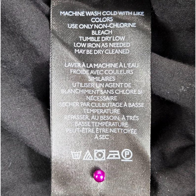 Puede incluir: Una etiqueta de ropa negra con texto blanco que detalla las instrucciones de lavado. La etiqueta incluye instrucciones en ingl&eacute;s y franc&eacute;s, con s&iacute;mbolos para lavar a m&aacute;quina, blanquear, secar y planchar. Una peque&ntilde;a cuenta rosa brillante est&aacute; unida a la etiqueta.