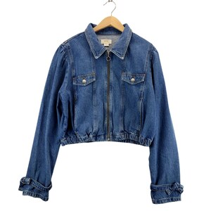 May include: A blue denim jacket with a zip-up front, two chest pockets, and elasticated waist and cuffs. The jacket is hanging on a wooden hanger. The sleeves have a unique design with a bow detail at the cuffs. The jacket is a medium wash denim.