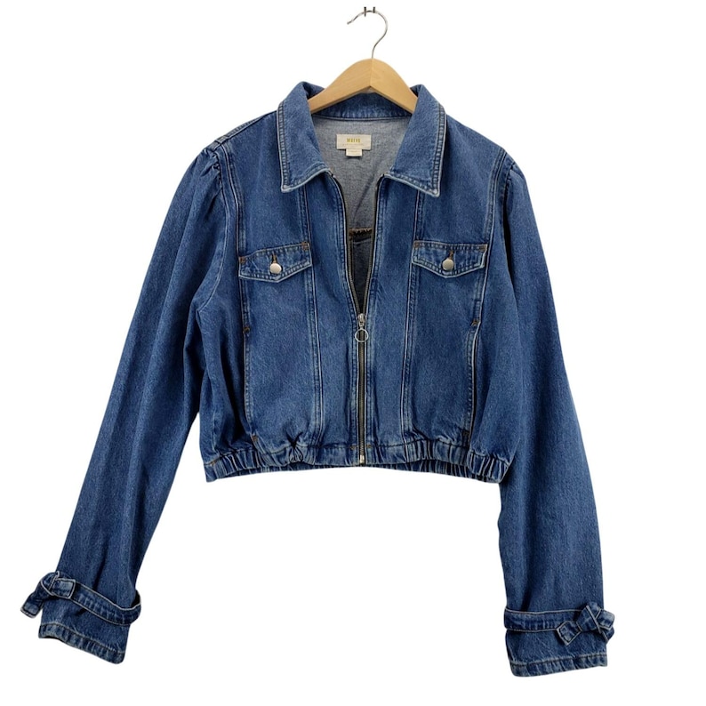 May include: A medium-wash blue denim jacket with a zip-up front, two chest pockets, and an elasticated waist. The long sleeves have bow details at the cuffs. The jacket is hanging on a wooden hanger.