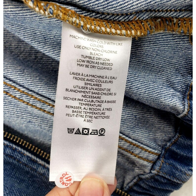 May include: A white clothing label with care instructions in English and French, attached to a denim garment. The label details washing, drying, and ironing instructions. The denim is blue.