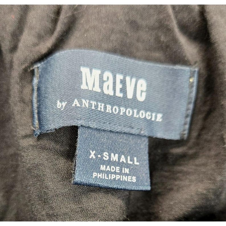 Puede incluir: Primer plano de una etiqueta de ropa azul marino con el nombre de la marca "Maeve by Anthropologie" en letras blancas. Debajo, una etiqueta m&aacute;s peque&ntilde;a que dice "X-SMALL" y "MADE IN PHILIPPINES".