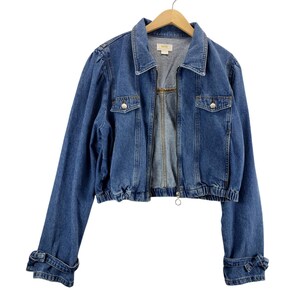 May include: A blue denim jacket with a zip-up front, two chest pockets, and buttoned cuffs. The jacket has a collared neckline and is hanging on a wooden hanger. The jacket's hemline is elasticated.